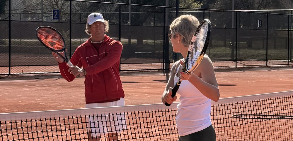 A coach and an adult player mirroring a stroke across the net
