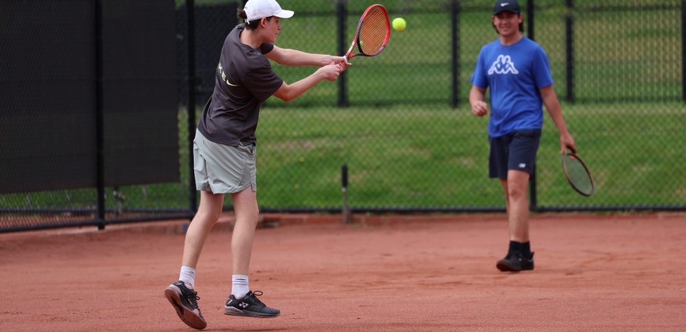 Two junior players trading shots on court, one hitting a high backhand