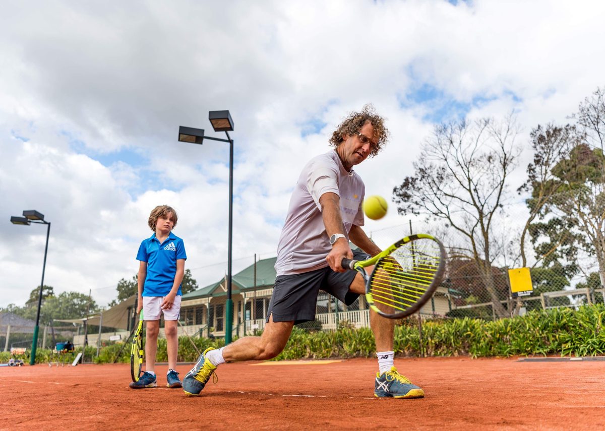 David Close demonstrating a low backhand on the clay court while a junior player watches